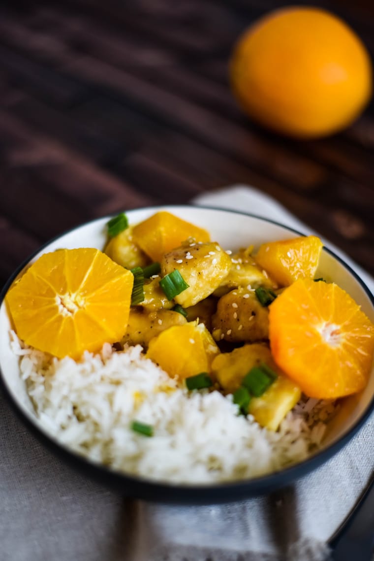 A white bowl of orange chicken with rice on a wooden table with an orange in the background 