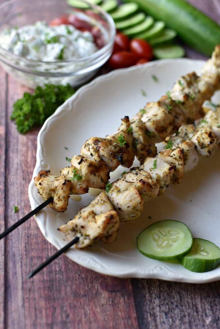 A white platter with skewers of Greek chicken kebabs and sliced cucumbers on a wooden table