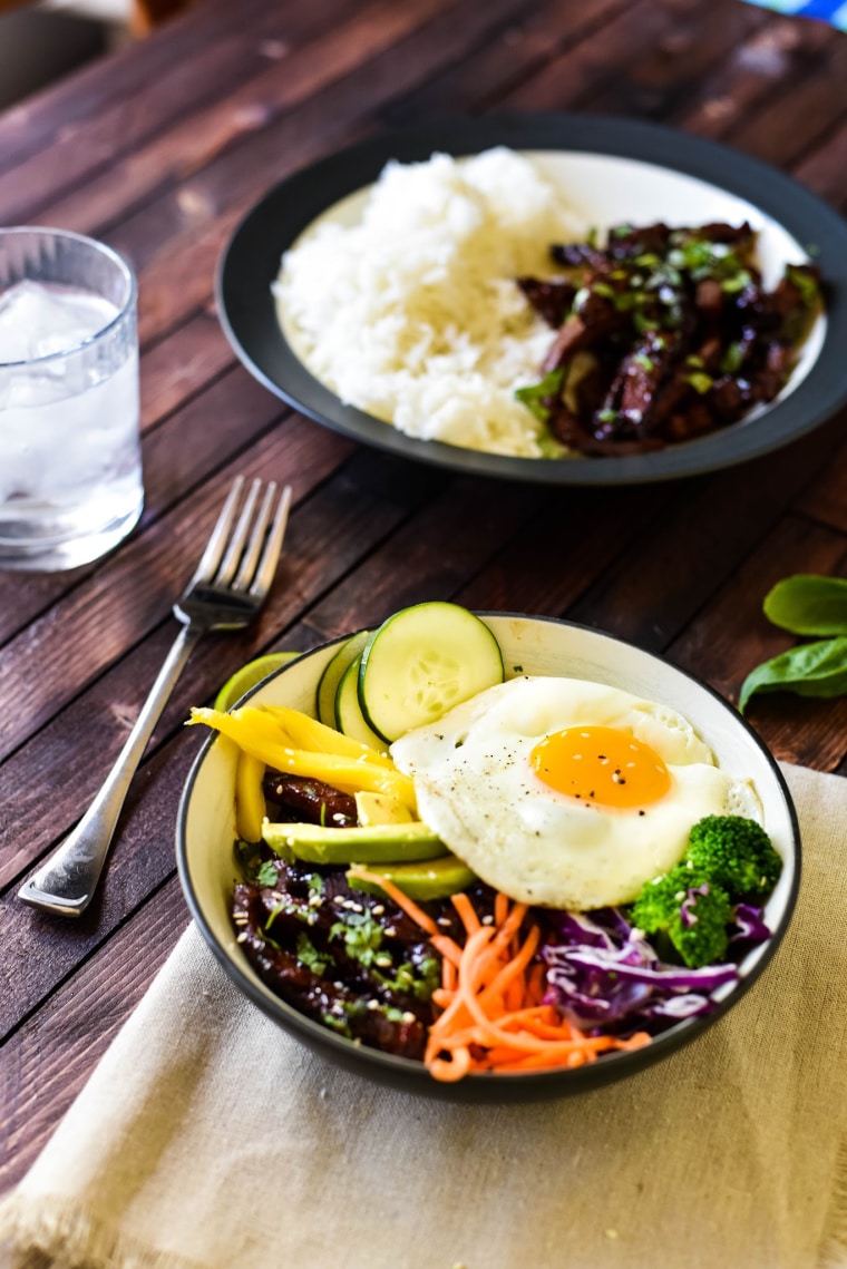 A plate of bulgogi and rice and a bowl of bulgogi with a fried egg on a wooden table with a fork and a glass of ice water