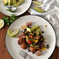 A plate of sausage sheet pan dinner on a wooden table with a plate of sliced avocado