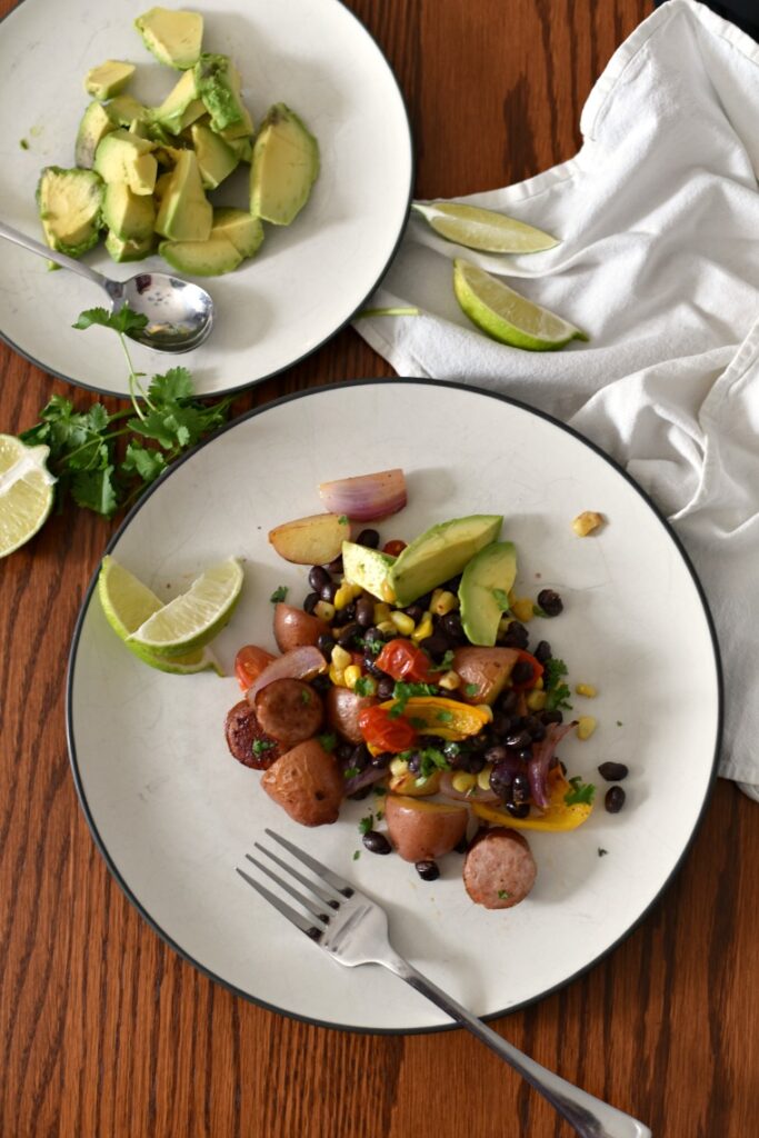 A plate of avocado slices and a plate of sausage sheet pan dinner on a wooden table with a white napkin