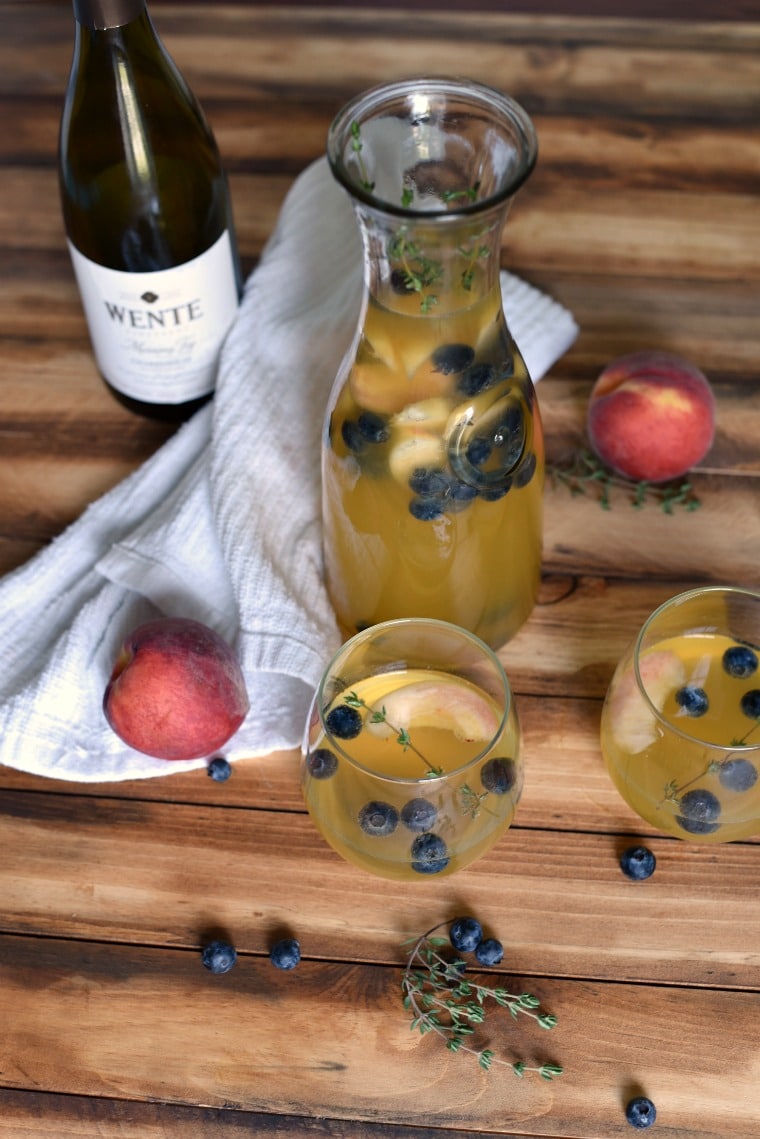 A karaffe and two glasses of summer sangria on a wooden table with a bottle of wine, a white towel, peaches, and blueberries