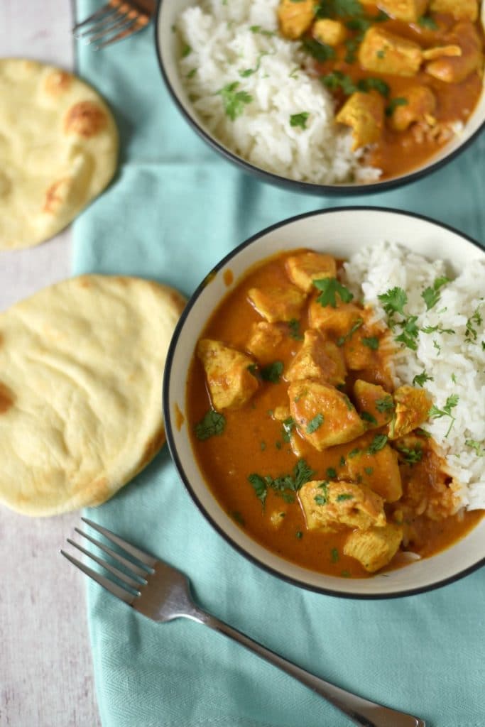 Two bowls of chicken tikka masala with rice and two naan breads on a blue napkin