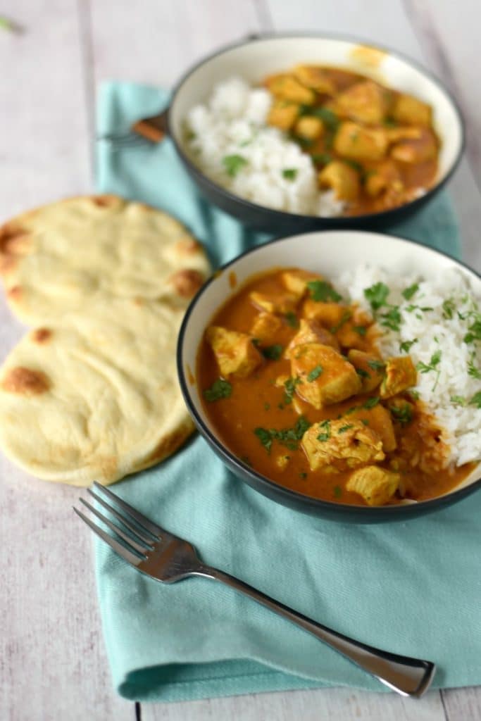 Two bowls of chicken tikka masala with rice and naan on a white wooden table with a blue napkin and silver fork