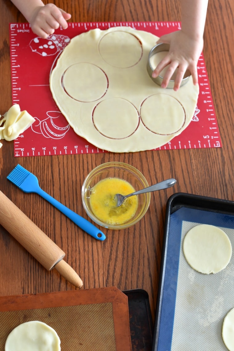 a child cutting out circles of dough with a round cookie cutter, sitting at a wooden table 