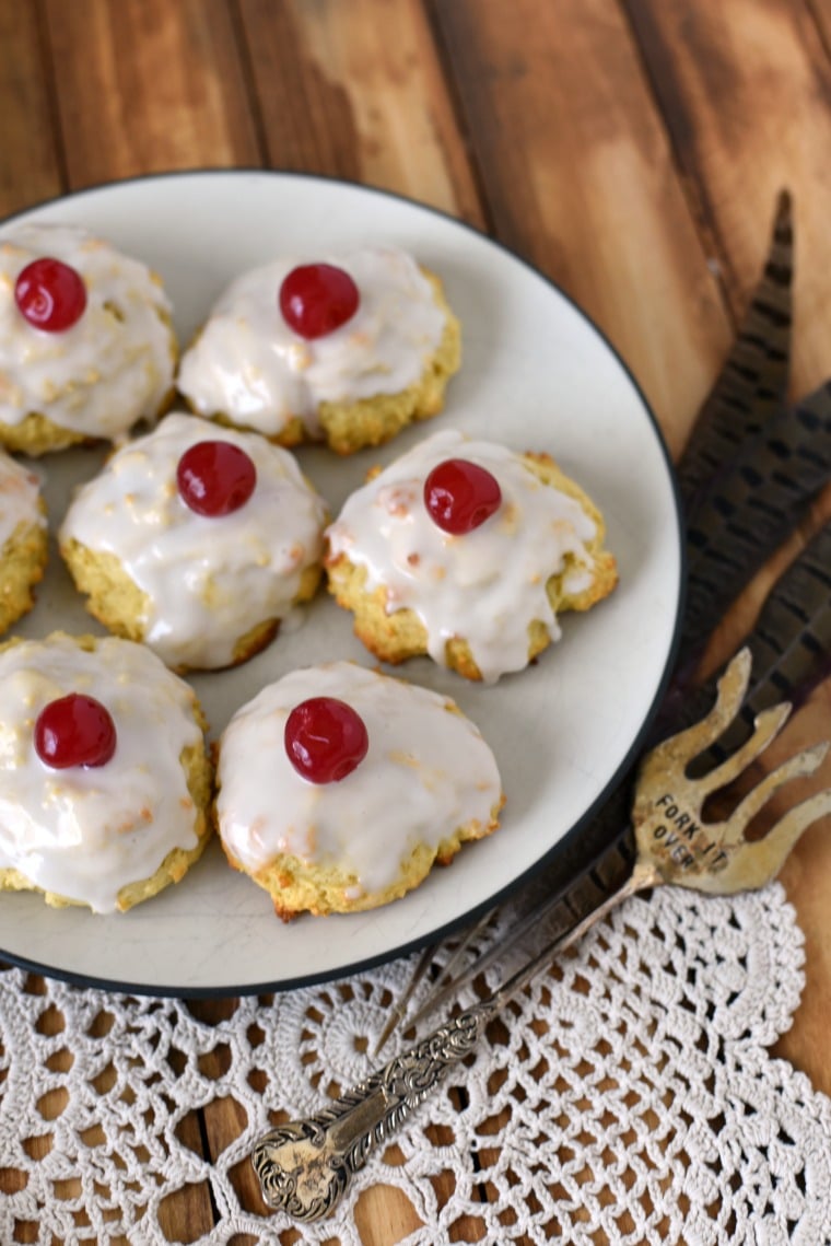 A white plate of empire biscuits on a wooden table with a white doilie and a silver serving fork