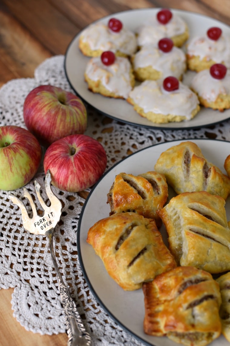 A white doilie on a wooden table with apples, a silver fork, a plate of empire biscuits, and a plate of sausage rolls