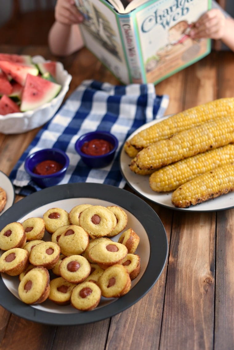 A child reading the book "Charlotte's Web" at a table with plates of watermelon, sweet corn, corndog muffins, and ketchup