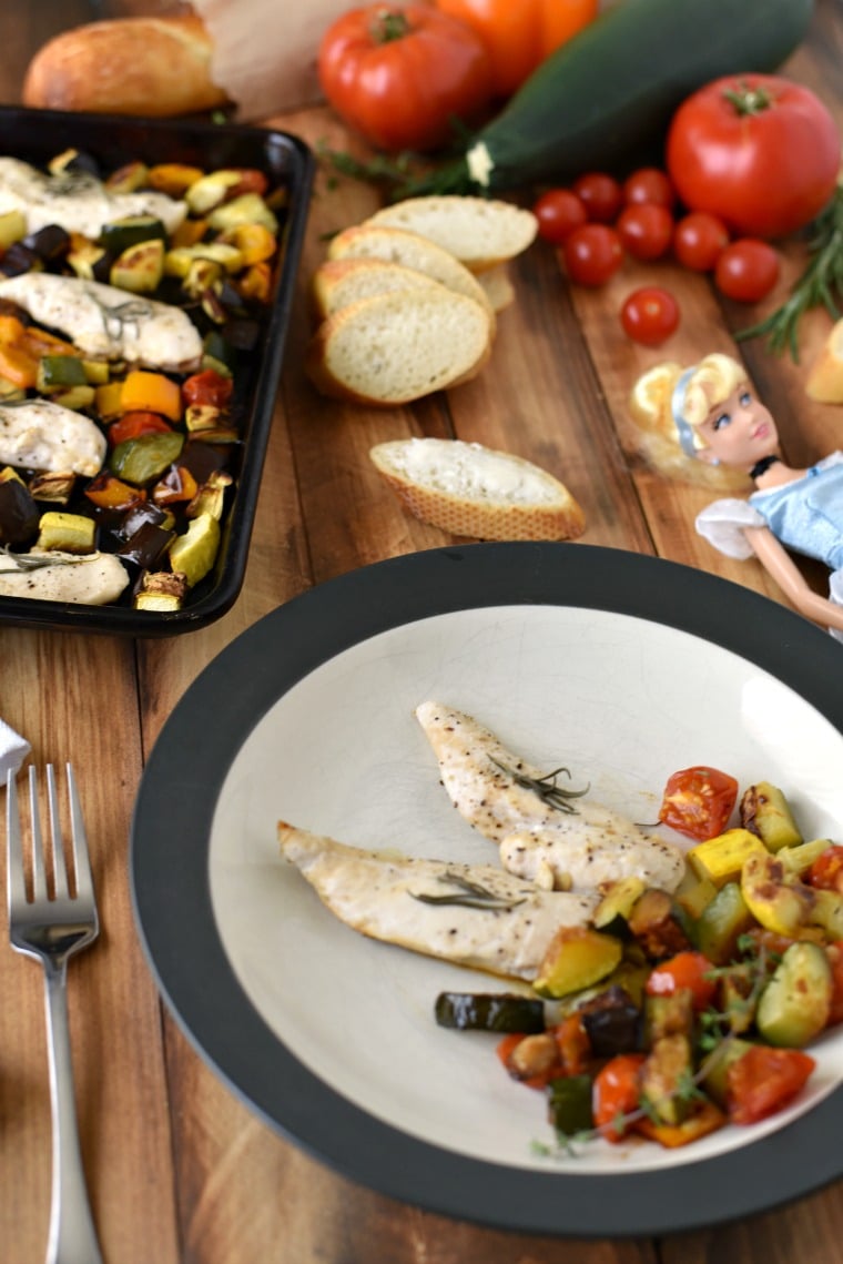 A sheet pan and plate of ratatouille and rosemary chicken on a table with sliced bread and a Cinderella doll