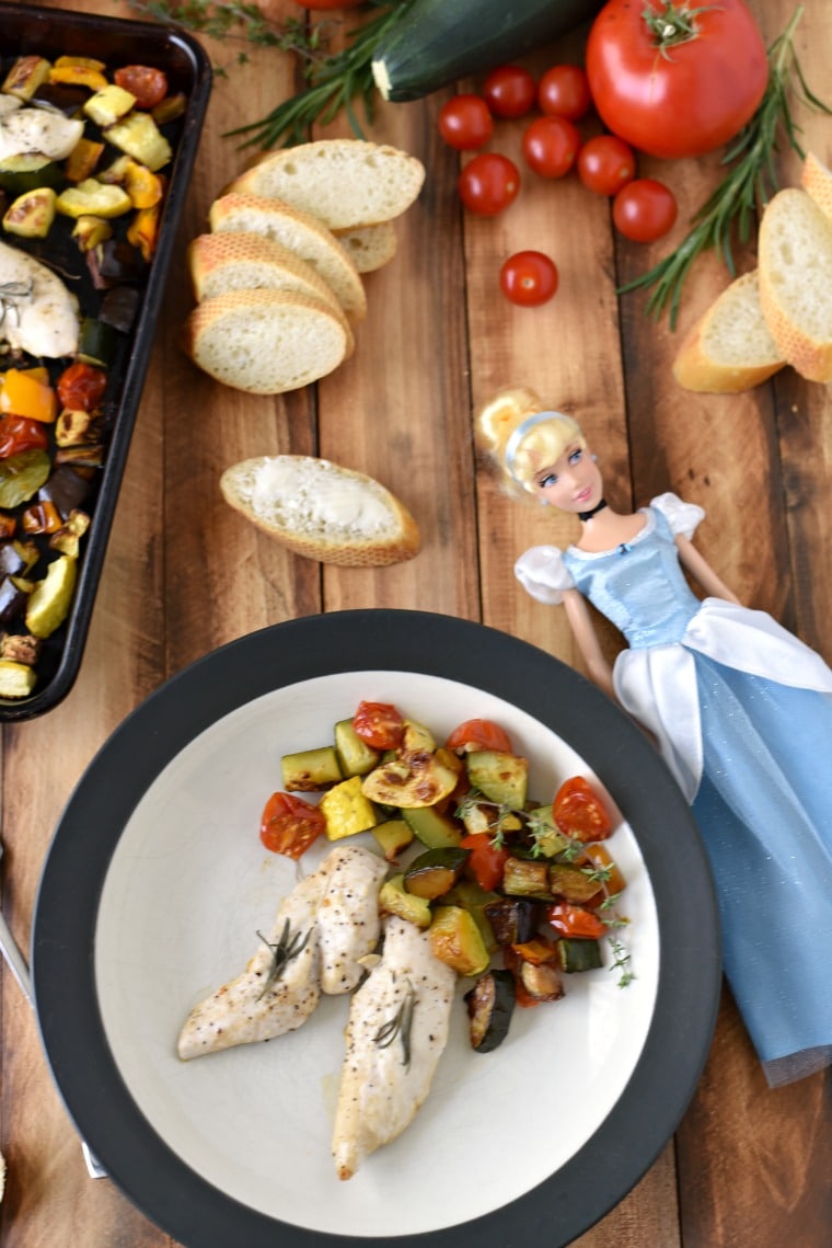 a Cinderella doll on a wooden table with sliced bread and a plate of ratatouille and rosemary chicken