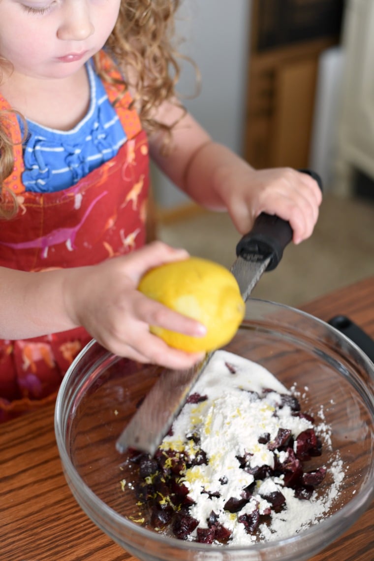 A child in a dinosaur apron zesting a lemon over a bowl of ingredients 