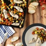 A sheet pan and a white plate on a wooden table, both with ratatouille and rosemary chicken
