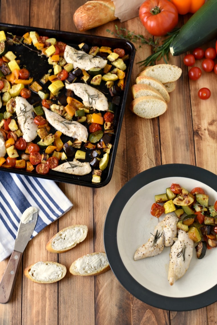 A sheet pan and a white plate on a wooden table, both with ratatouille and rosemary chicken