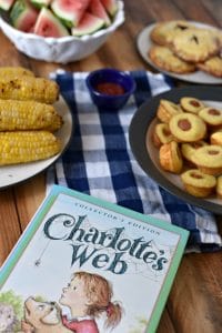 The book Charlotte\'s Web on a wooden table with a picnic on a blue checkered tablecloth behind