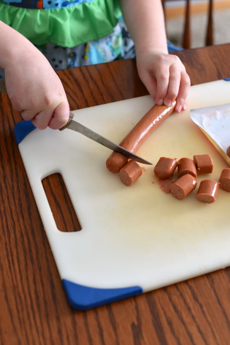 A small child slicing hot dogs on a white cutting board with a knife
