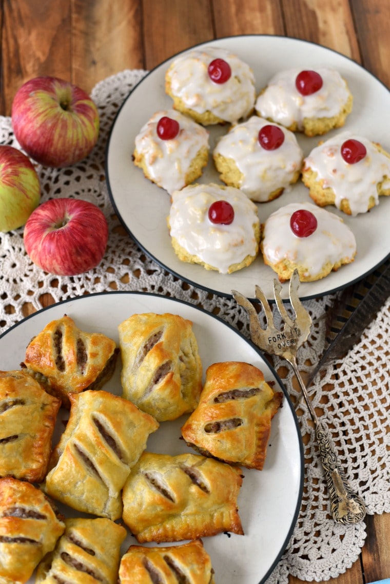 Two white plates on a white doilie with apples and a silver serving fork. One plate has sausage rolls and the other plate has empire biscuits