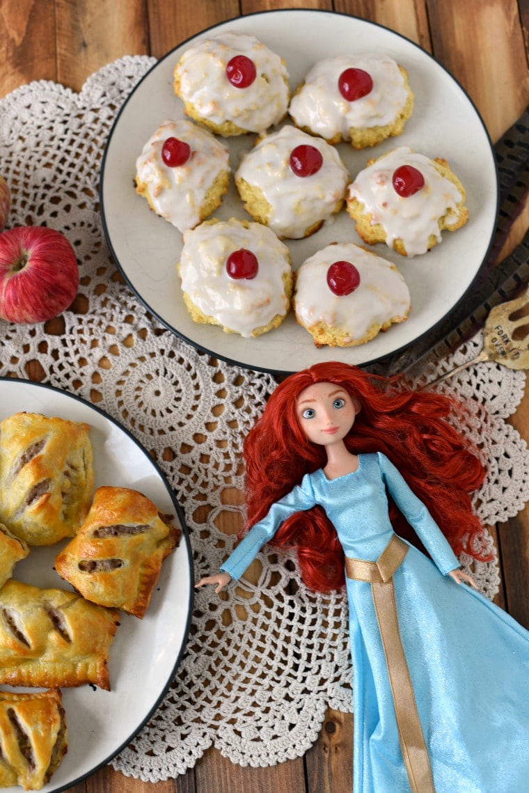 A plate of sausage rolls and a plate of empire biscuits on a white doilie with a Merida doll and a red apple