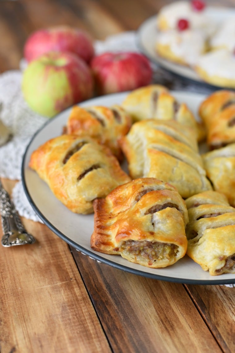 A plate of sausage rolls on a wooden table with apples in the background