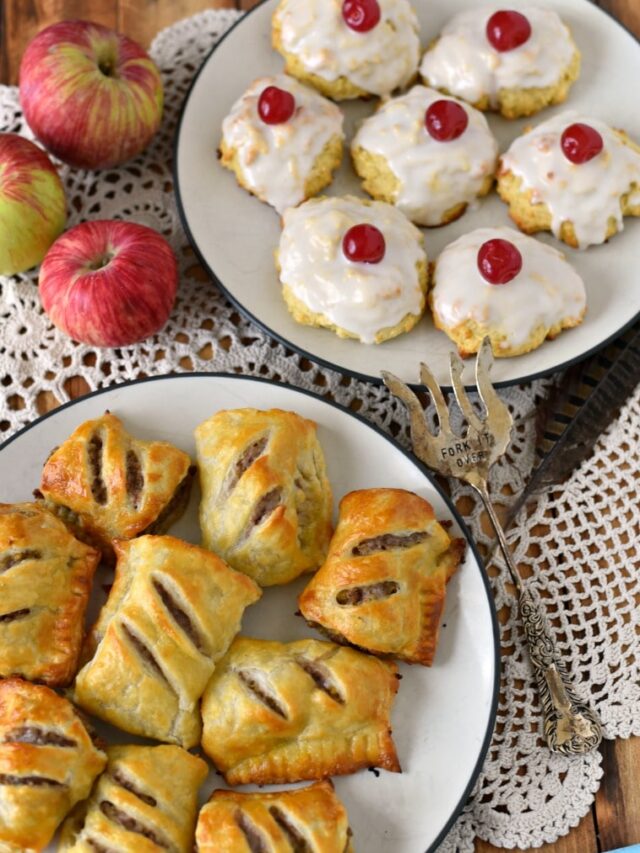 Two white plates on a white doilie with apples and a silver serving fork. One plate has sausage rolls and the other plate has empire biscuits