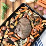 A wooden table with bundled herbs, carrots, potatoes, and a sheet pan dinner of roasted veggies and roast beef