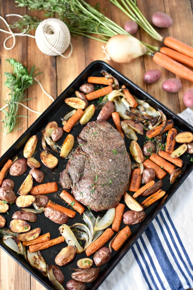A wooden table with bundled herbs, carrots, potatoes, and a sheet pan dinner of roasted veggies and roast beef