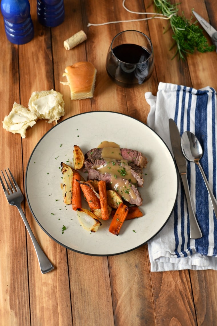 A wooden table set with a blue and white napkin, silverware, a white plate of roast beef sheet pan dinner, and a buttered dinner roll 