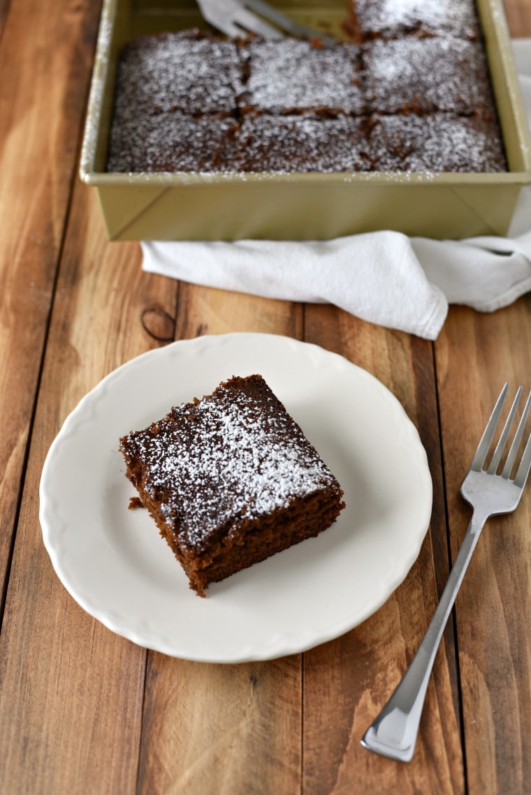 A pan of gingerbread cake and a plate with a slice of gingerbread cake, both on a wooden table