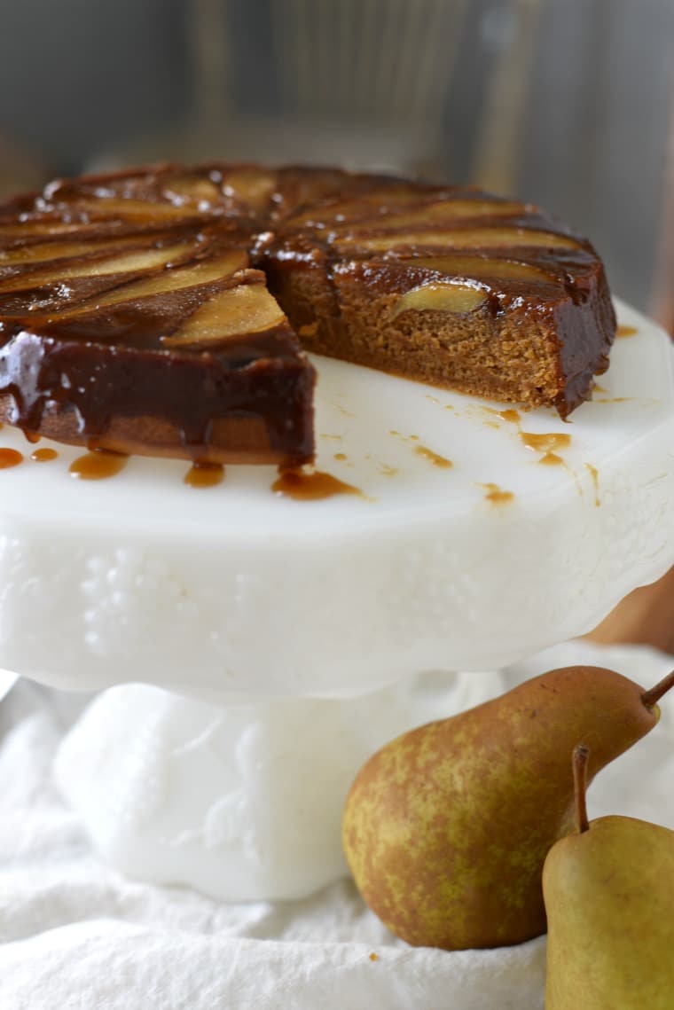 Pear gingerbread cake with a slice missing on a white cake platter with whole pears in the foreground