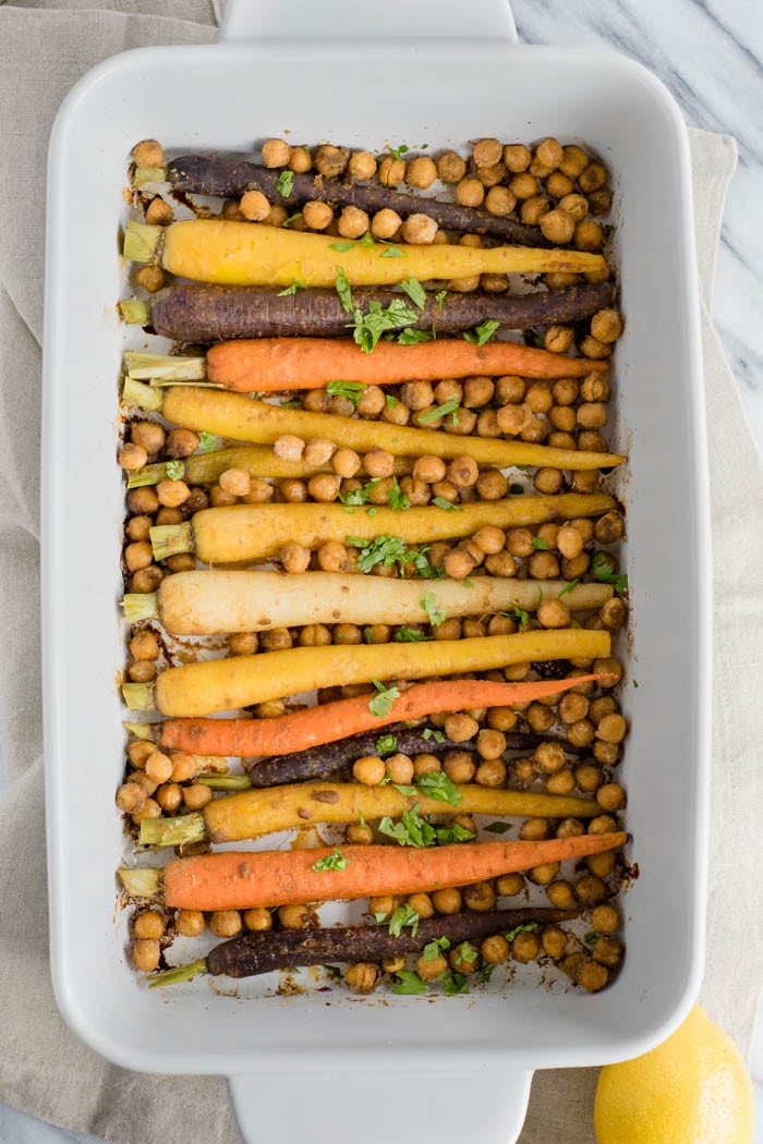 A white baking dish filled with chickpeas, whole rainbow carrots, and sprinkled with green herbs