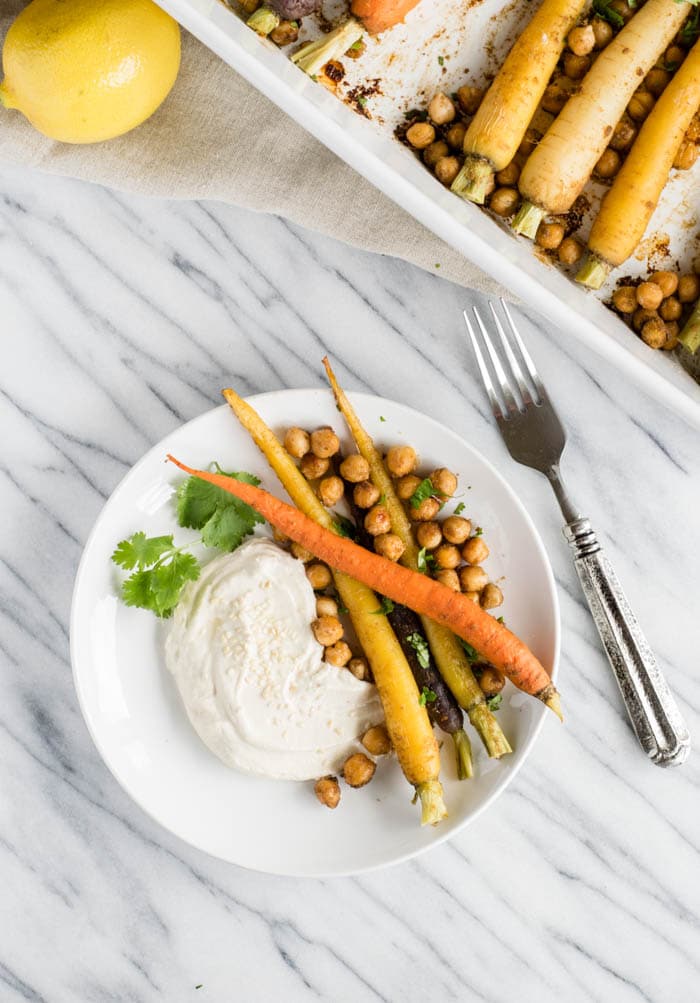A white marble countertop with a white baking dish an white plate, both with whole rainbow carrots, chickpeas, and cilantro. A silver fork between