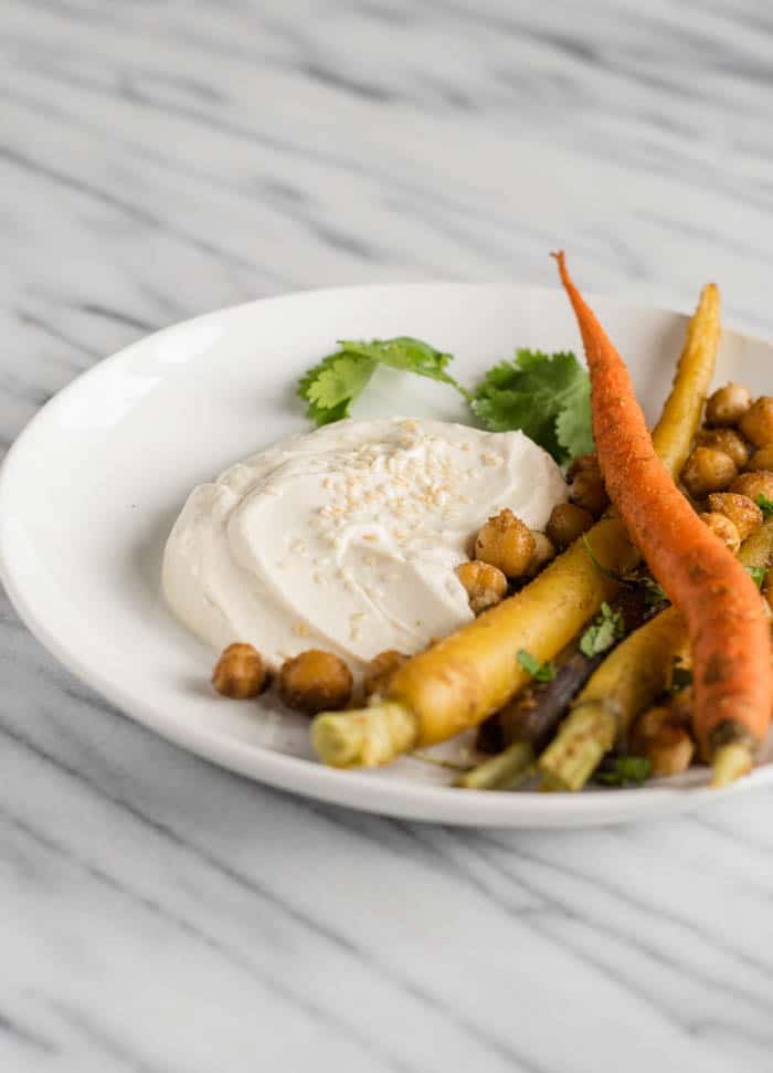 A white plate on a white marble countertop with tahini yogurt next to whole rainbow carrots and chickpeas