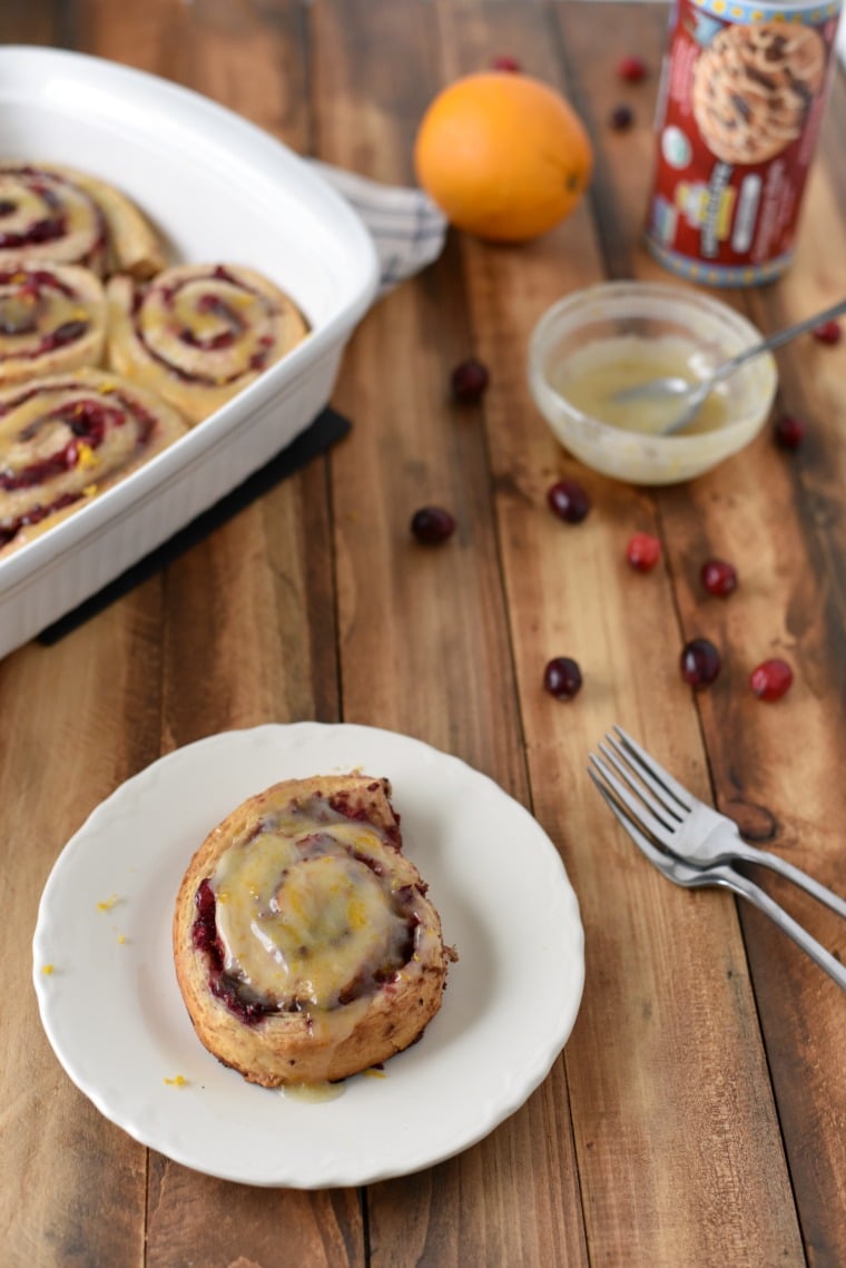 A plate with a cranberry orange cinnamon roll on a wooden table, with forks, cranberries, icing bowl, and baking dish also on the table