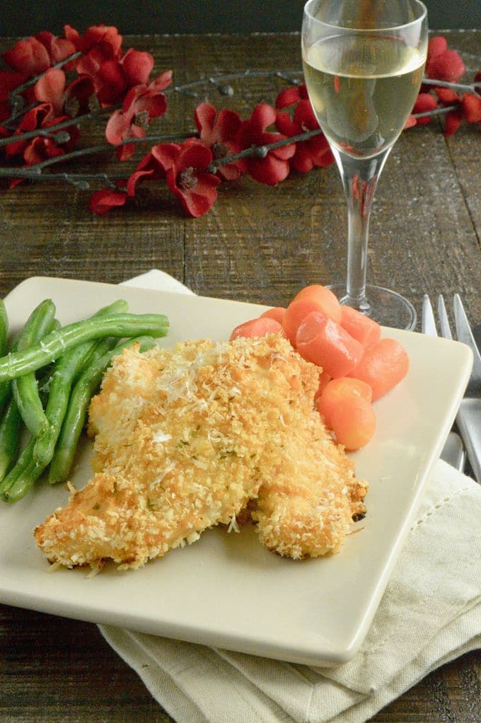 A white plate on a white napkin with green beans, breaded chicken tenders, carrots sticks, and a wine glass. All on a wooden table. 