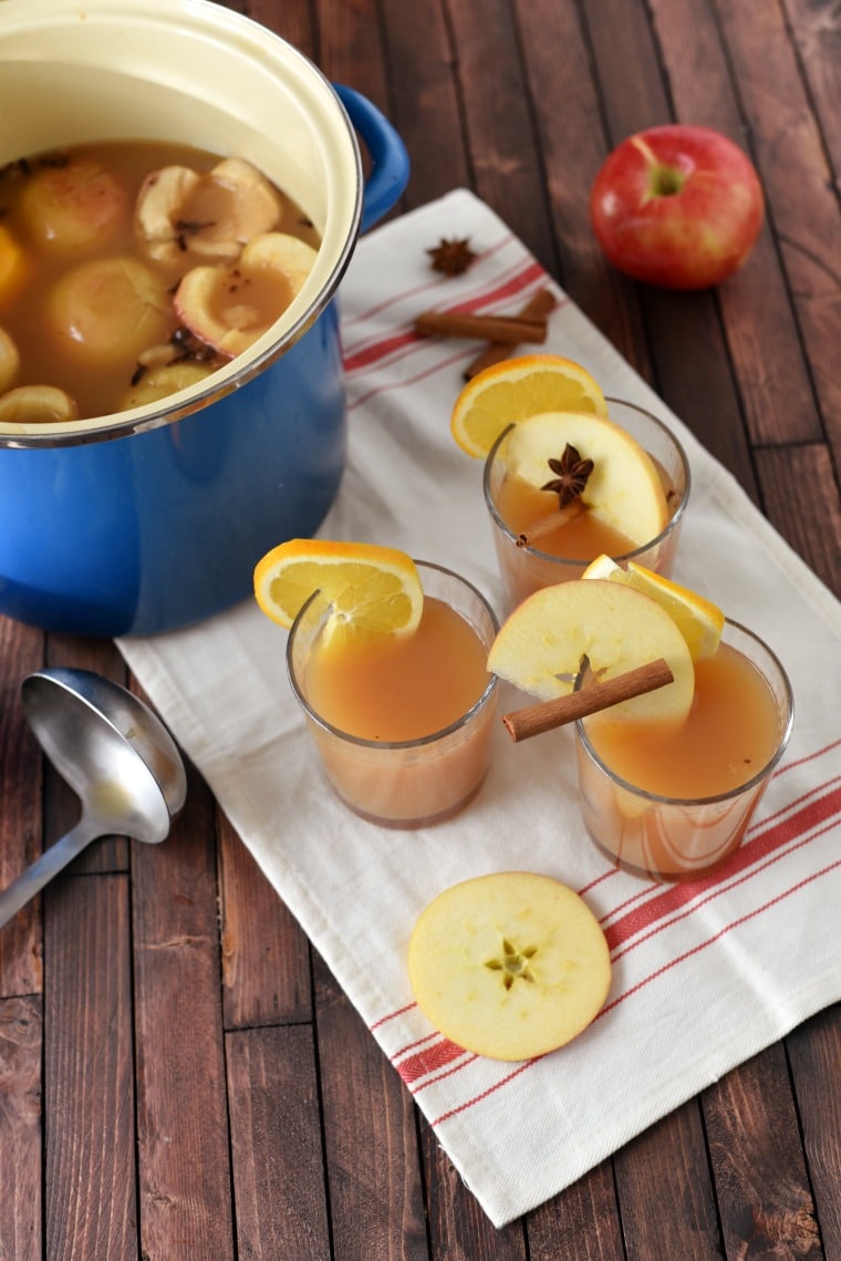 Three glasses of apple cider wassail, and a blue pot with apple cider wassail inside. All on a wooden table and white and red napkin. A silver ladel is on the table beside