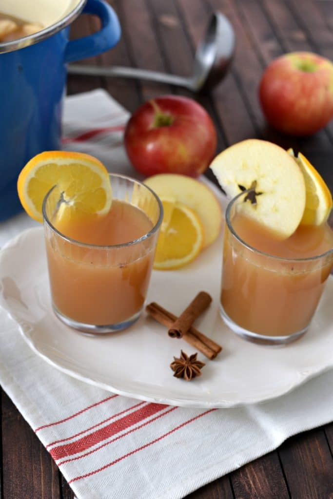 Two glasses of apple cider wassail on a white plate with crossed cinnamon sticks, star anise, and apples around 