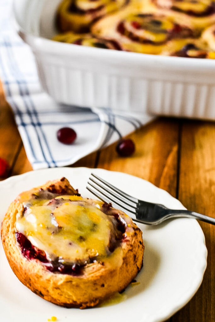 cinnamon roll with cranberry and orange on white plate with fork, with baking more in background