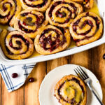 overhead view of baking tray with orange cinnamon rolls with cranberries and one on plate
