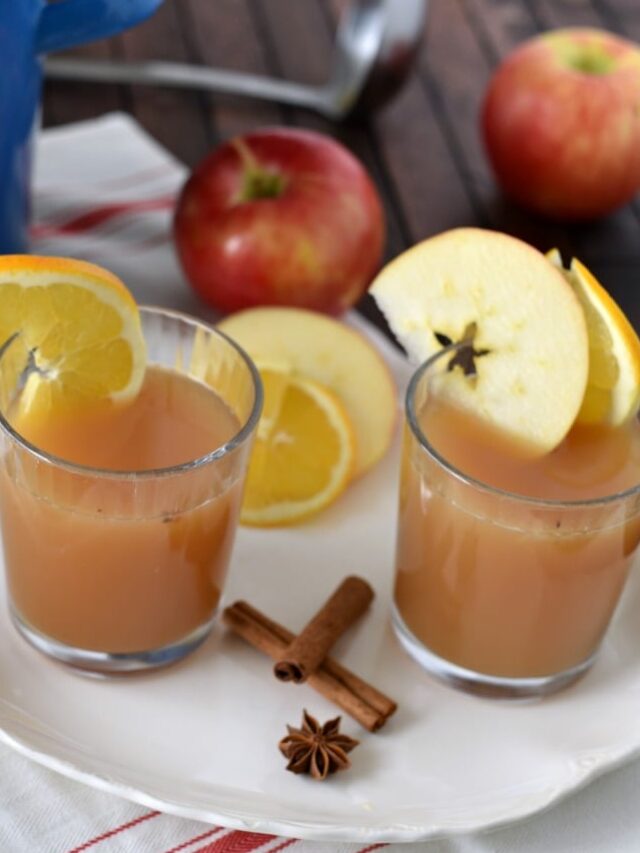 Two glasses of apple cider wassail on a white plate with crossed cinnamon sticks, star anise, and apples around