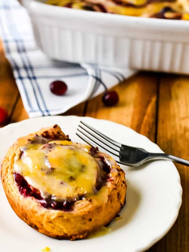 cinnamon roll with cranberry and orange on white plate with fork, with baking more in background