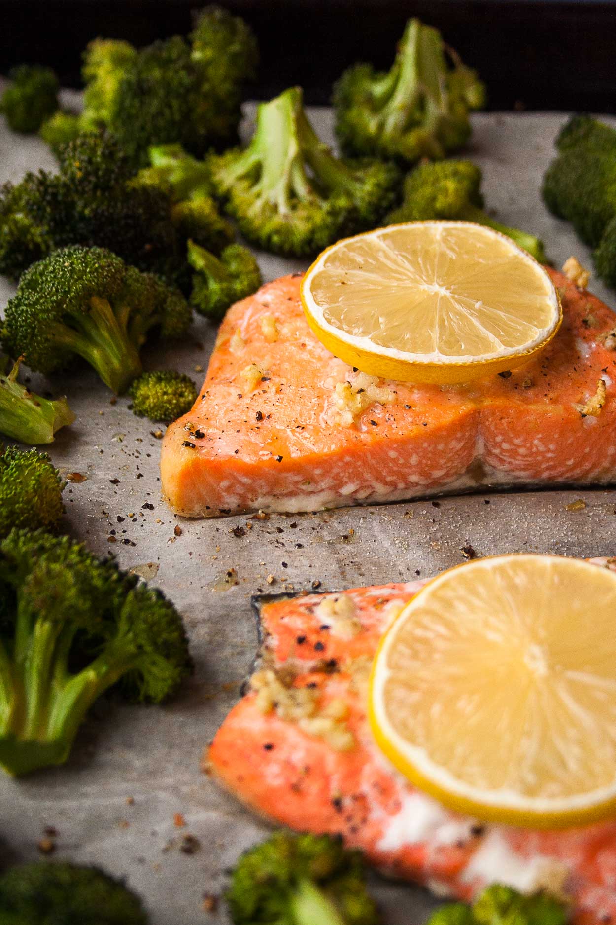 Two cooked salmon fillets covered in lemon slices next to roasted broccoli on top of parchment paper 