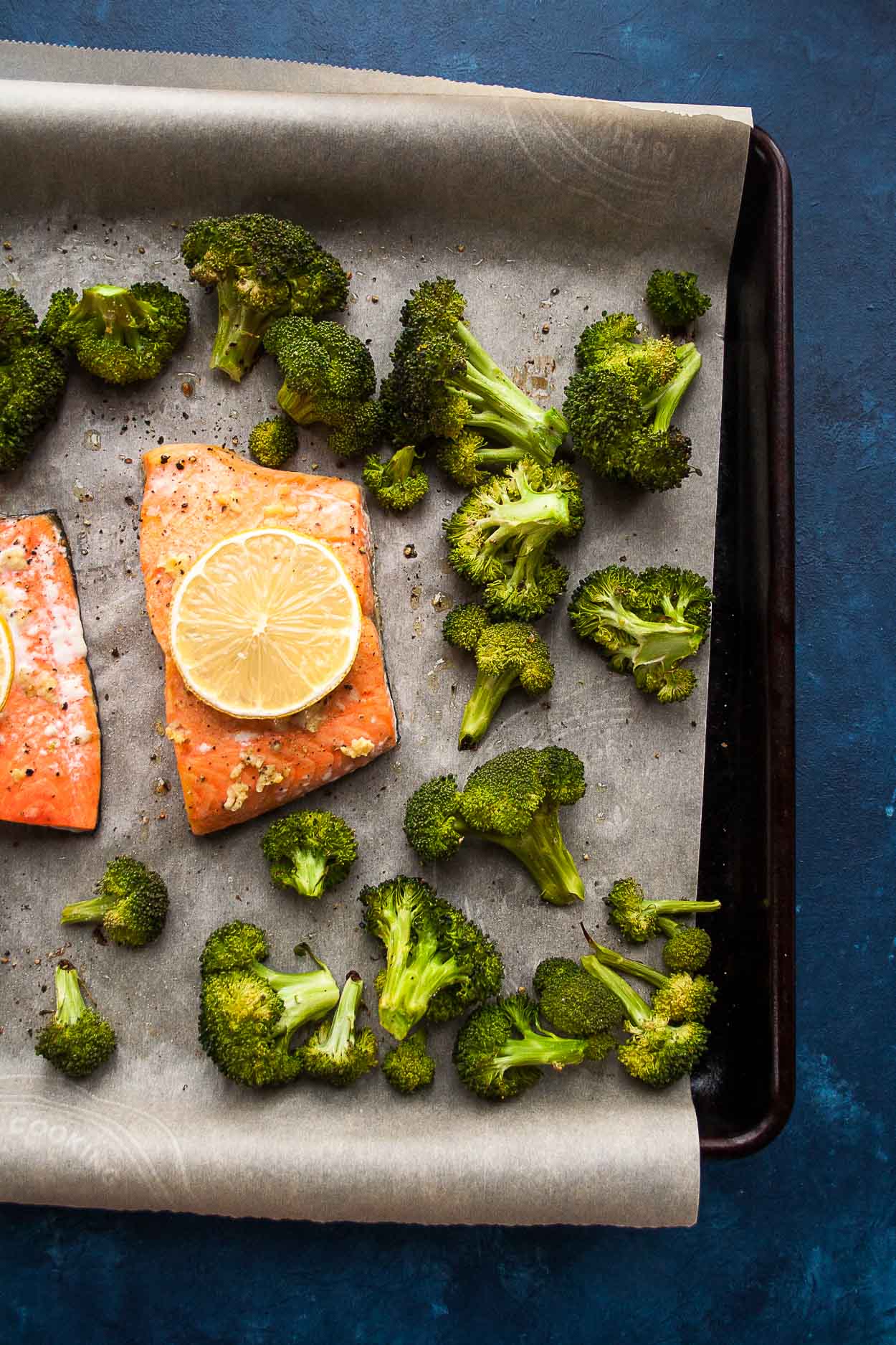 A baking sheet with parchment paper, roasted broccoli, two salmon fillets covered with lemon slices, all on a blue table 