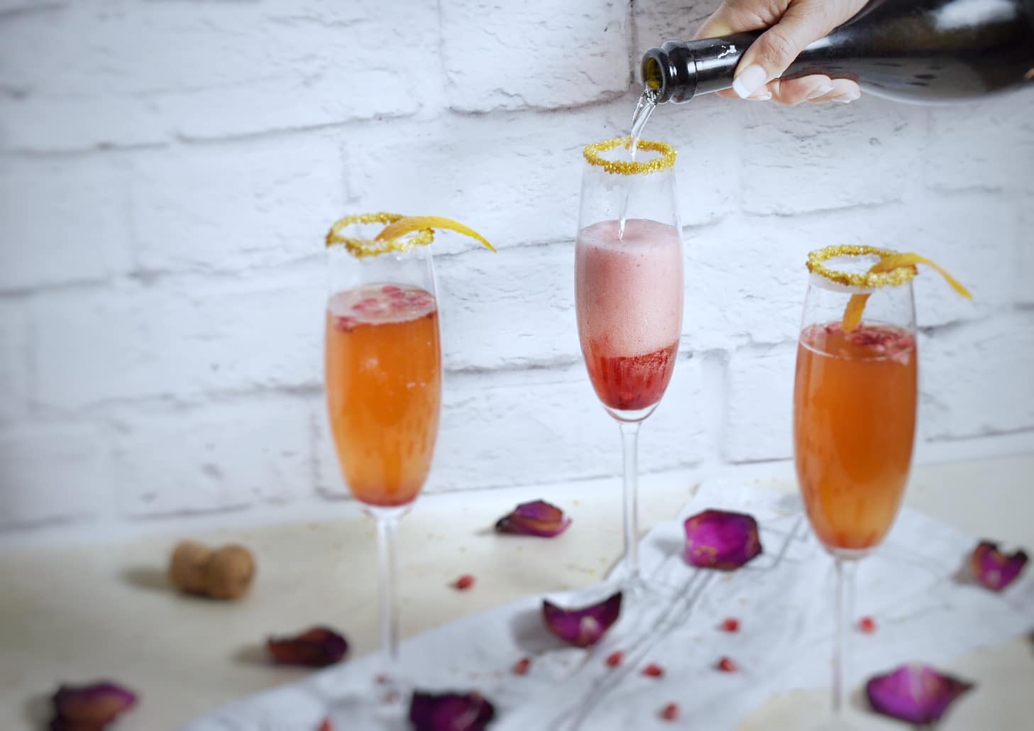 A Caucasian hand pouring champagne from a bottle into the middle of three champagne flutes filled with orange pomegranate champagne cocktails 