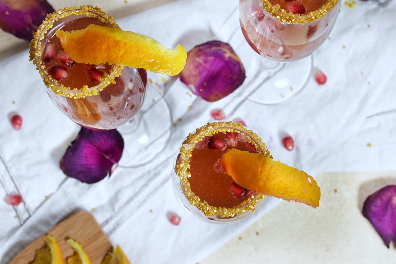A top down view of three champagne flutes, rimmed in sugar, on a white counter, filled with orange pomegranate cocktail and and orange peel garnish