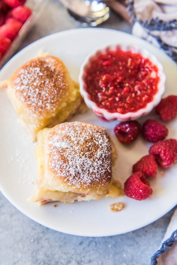 A white plate with two monte cristo sliders, raspberries, and a small bowl of red jam