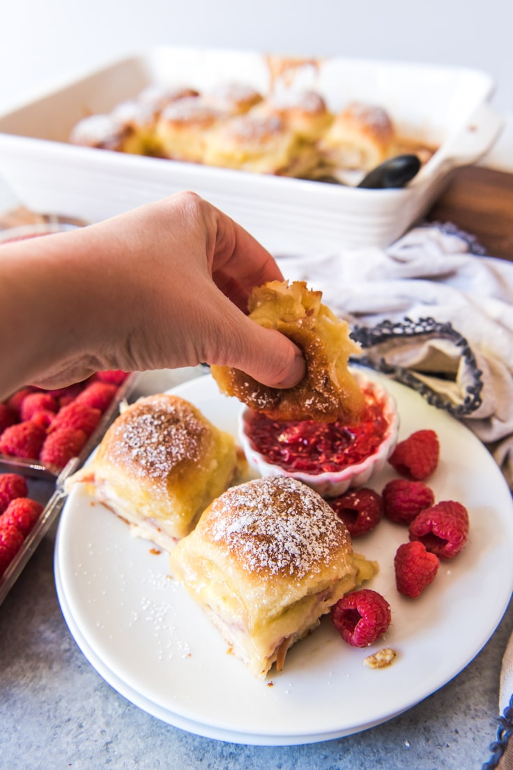 A Caucasian hand holding a monte cristo slider and dipping it into a small bowl of raspberry jam that is on a plate with two other sliders and raspberries 