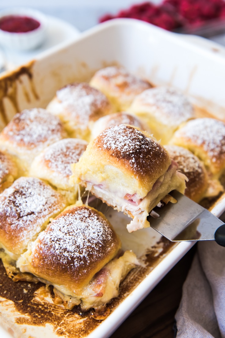 A silver spatula lifting a monte cristo slider out of its baking dish that has 11 other sliders inside still