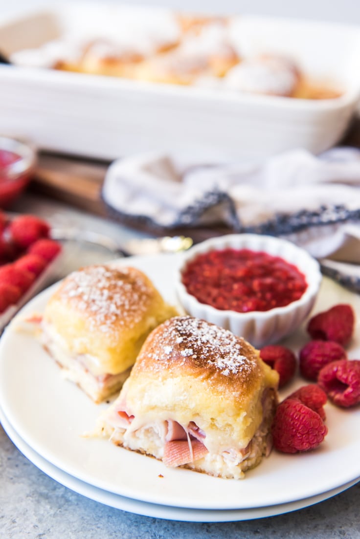 A plate with two monte cristo sliders dusted in powered sugar, 6 raspberries, and a small bowl of raspberry jam on a wooden table, a white baking dish behind