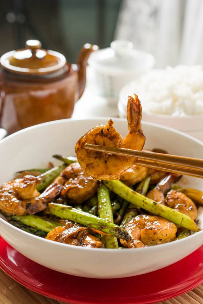 A pair of chopsticks holding a cooked shrimp over a white bowl of stir fried shrimp and asparagus 