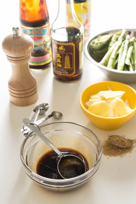 A salt shaker, a bottle of soy sauce, a bowl with asparagus, a bowl of chopped ginger, some black pepper in a silver spoon, and a bowl of soy sauce with a measuring spoon inside