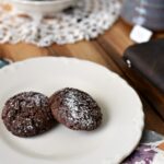 Two chocolate cookies dusted with powdered sugar sitting on a white plate on a wooden table with a book and ;ace doily next to it